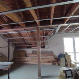 Poddasze w trakcie prac: widoczne drewniane belki stropowe, metalowa konstrukcja pod przyszły sufit, stosy materiałów izolacyjnych i desek pod ścianą, przygotowane do dalszych prac wykończeniowych.