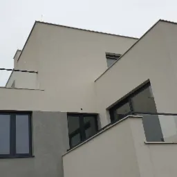 Nowoczesny budynek z balkonami zabezpieczonymi szklanymi balustradami z czarnymi poręczami, widok z dołu na tle pochmurnego nieba.