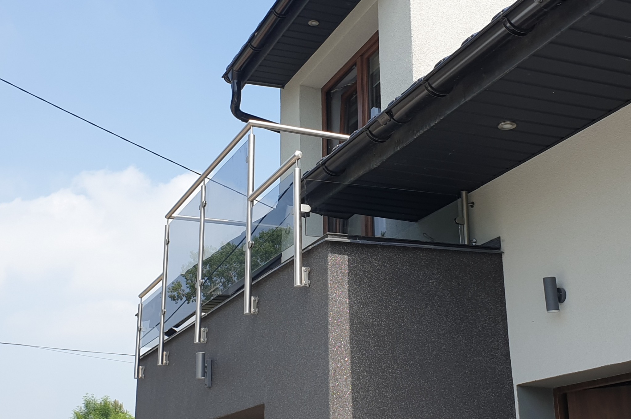 Szklana balustrada balkonowa z nierdzewnymi słupkami, widok z dołu na tle błękitnego nieba z chmurami.