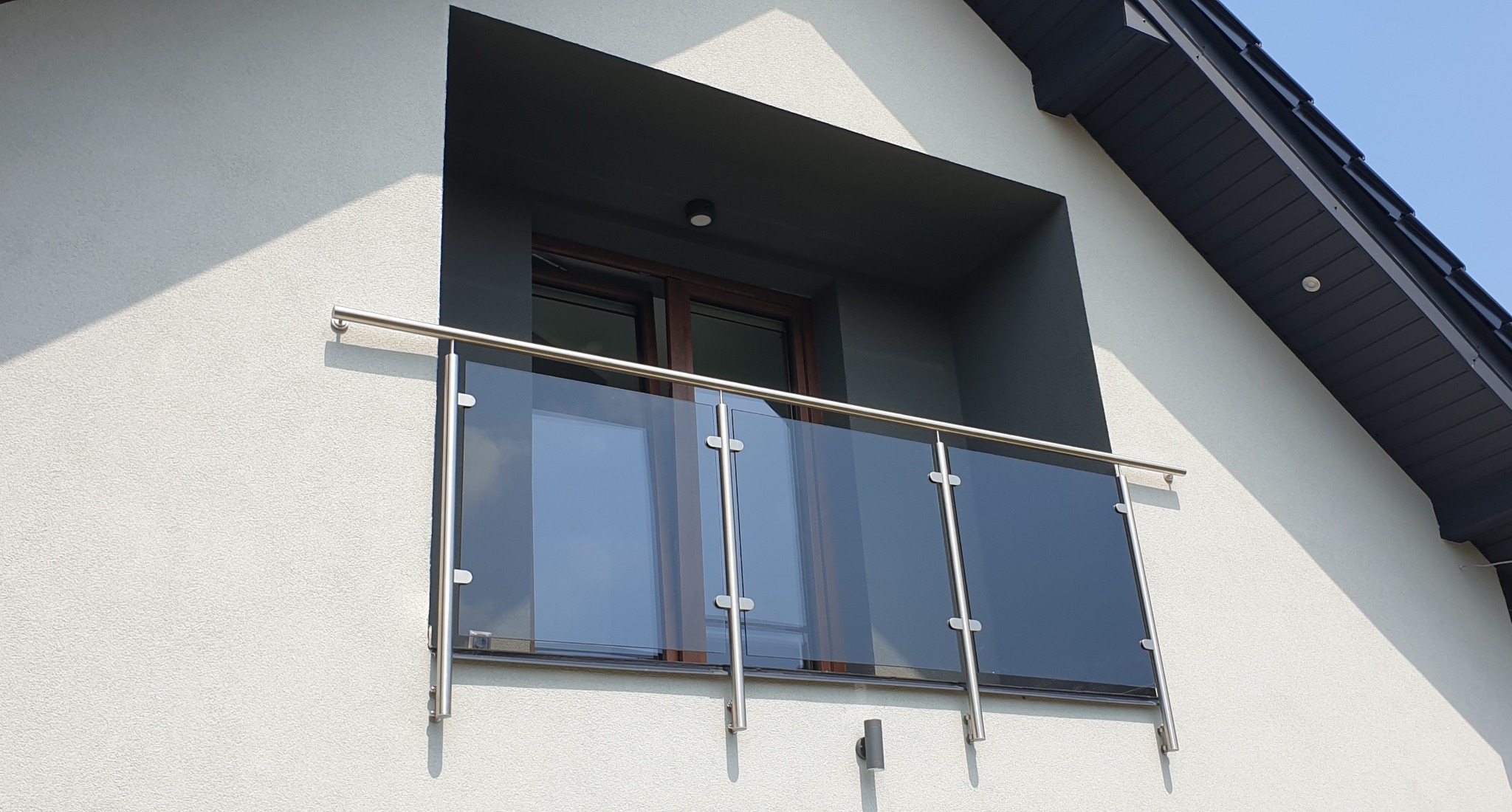 Balkon z nowoczesną szklaną balustradą z elementami ze stali nierdzewnej, widok z dołu na jasną elewację budynku.