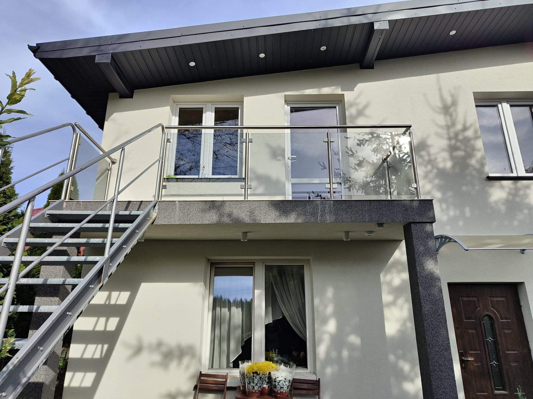 Zewnętrzne schody stalowe prowadzące na balkon z przeszkloną barierką w nowoczesnym domu z szarą elewacją.