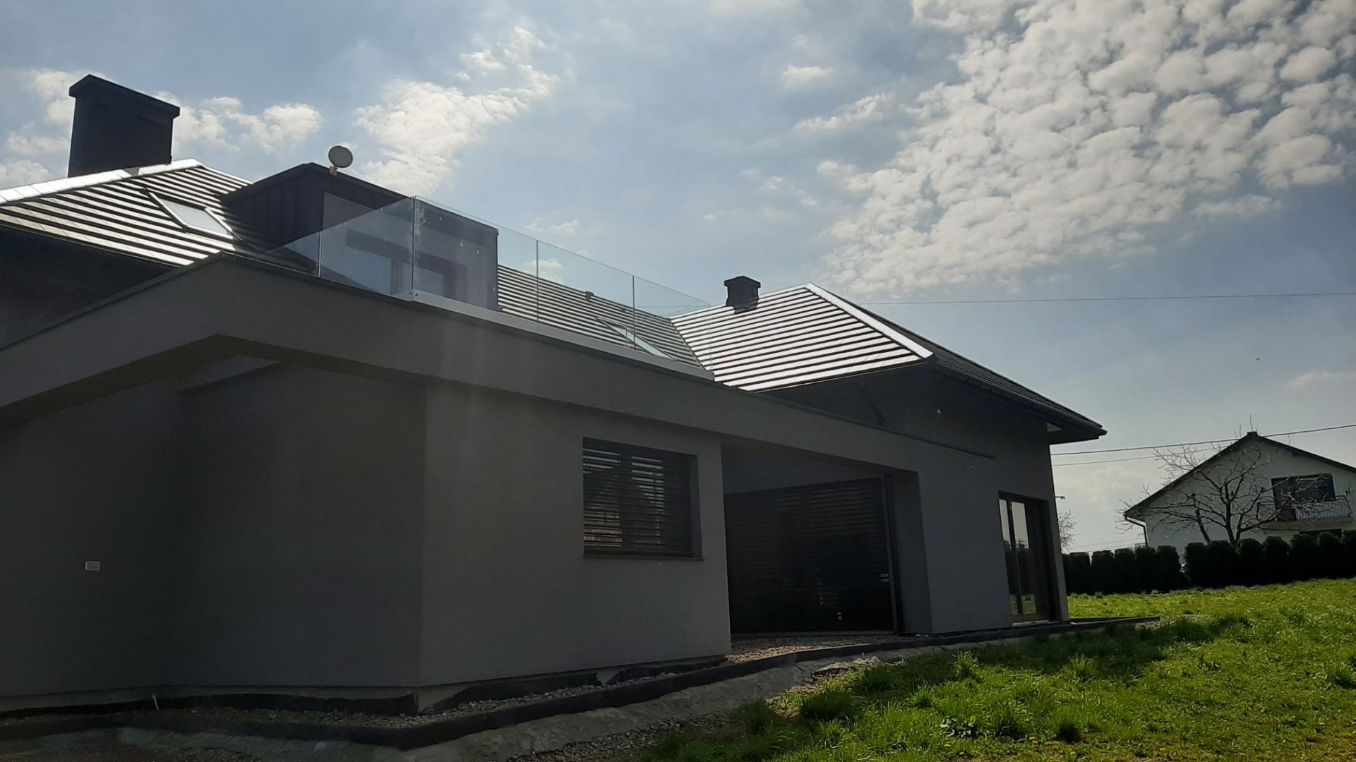Nowoczesny dom z szarą elewacją i dachem z blachy trapezowej, na dachu zamontowana szklana barierka balkonowa, widok z poziomu gruntu, w tle błękitne niebo z chmurami.