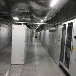 Długi korytarz z rzędami białych szaf elektrycznych, widoczne instalacje wentylacyjne pod sufitem i metalowy podest na podłodze.