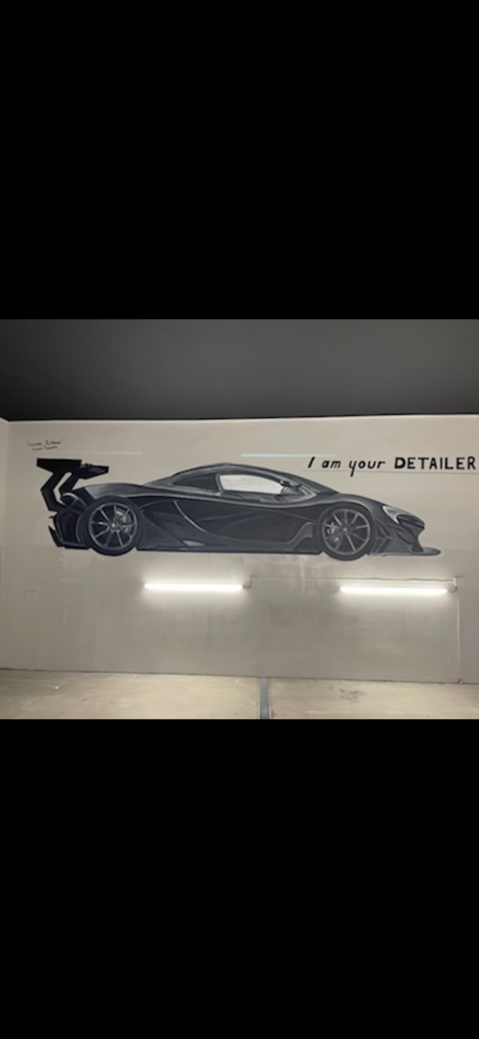 Czarno-biały mural przedstawiający sportowy samochód z napisem 'I am your DETAILER' na szarej ścianie, oświetlony dwoma lampami jarzeniowymi.