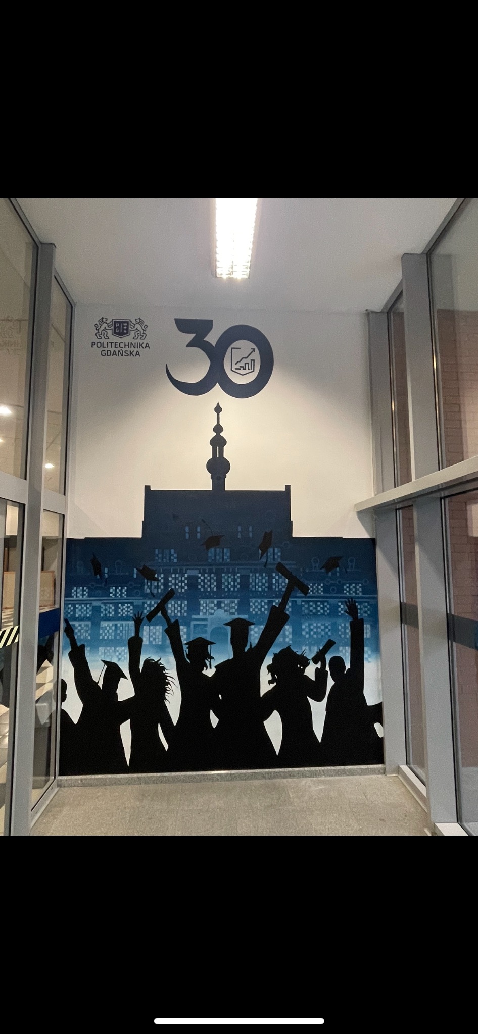 Malowidło ścienne w korytarzu uczelni z sylwetkami absolwentów rzucających birety na tle budynku Politechniki Gdańskiej i logo 30-lecia.