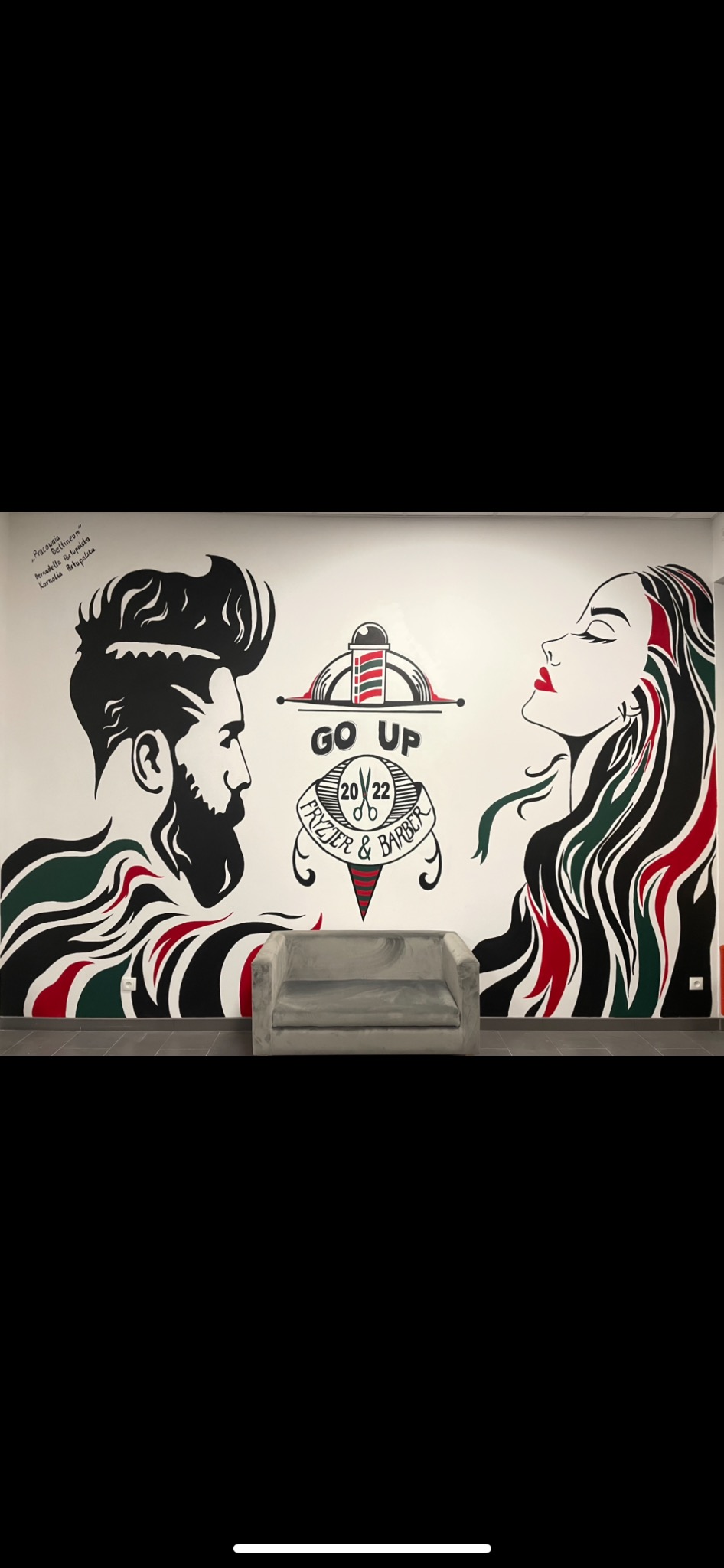 Ściana w salonie fryzjerskim z czarno-białym malunkiem przedstawiającym profil brodatego mężczyzny i kobiety z czerwonymi ustami, logo salonu 'Go Up Fryzjer & Barber' oraz szara sofa przed ścianą.