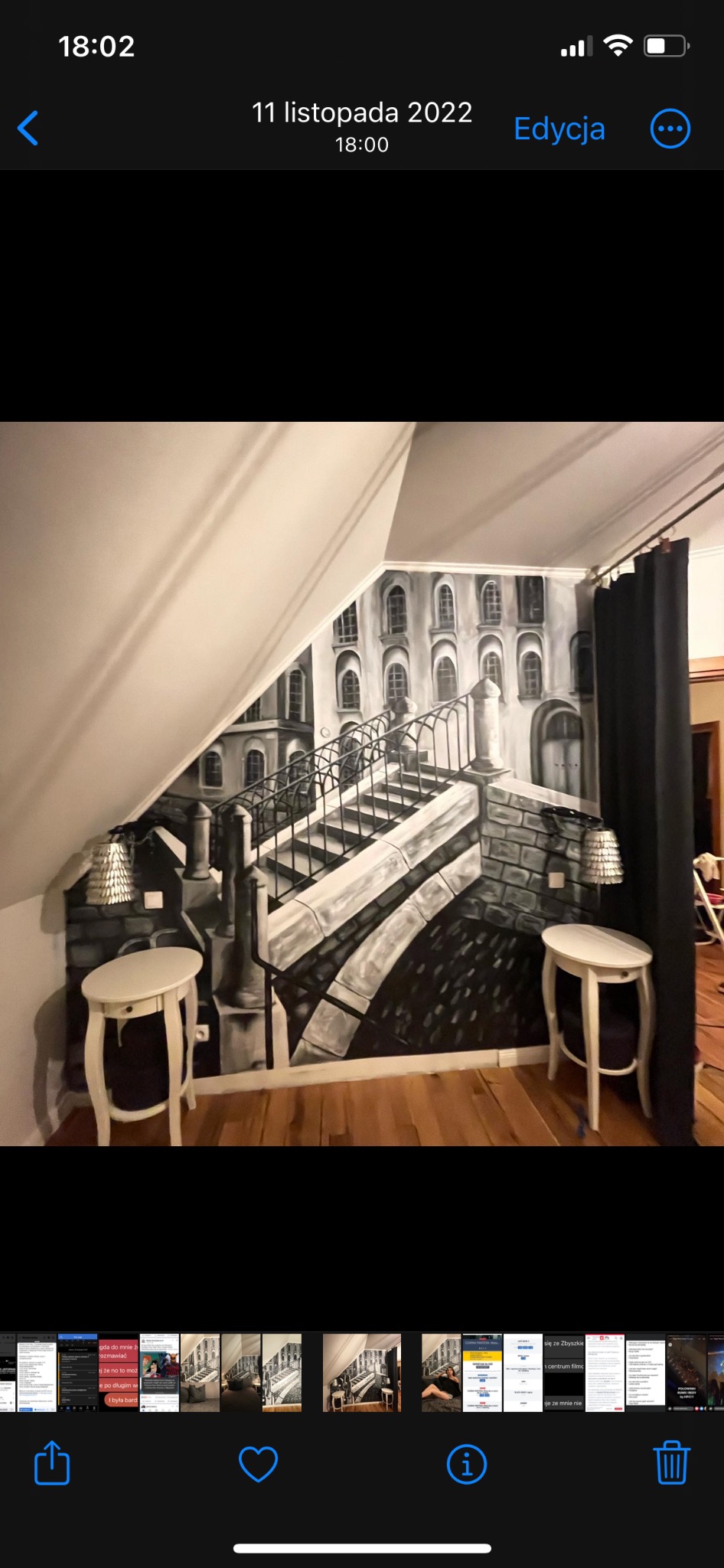 Czarno-biała iluzjonistyczna malowanka na ścianie pod skosem, przedstawiająca schody i budynki w stylu starej architektury miejskiej, z dwoma małymi białymi stolikami po bokach.