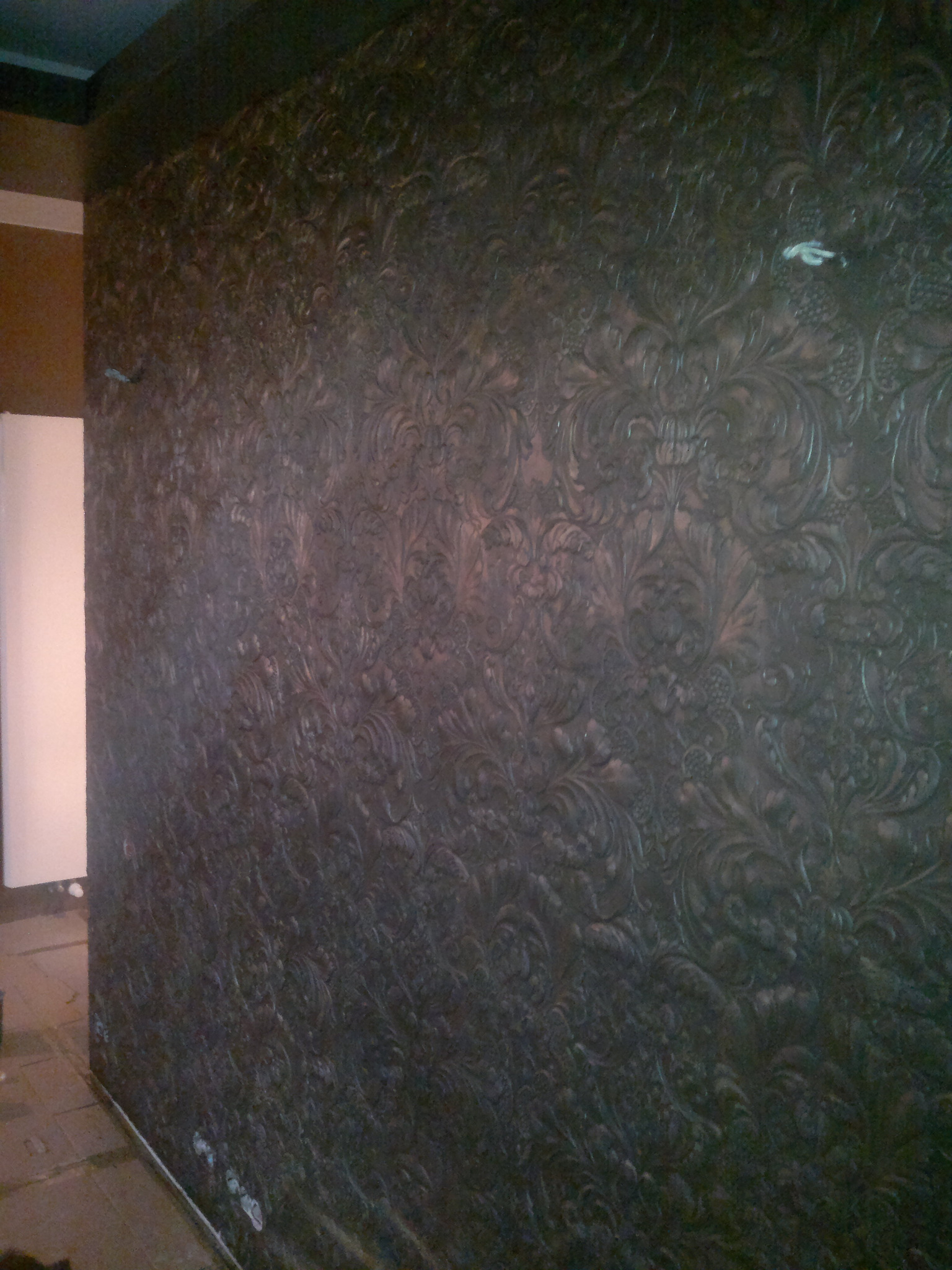Teksturowana ściana w ciemnych odcieniach brązu z wytłaczanym wzorem roślinnym, widoczny fragment sufitu i sąsiedniej ściany.