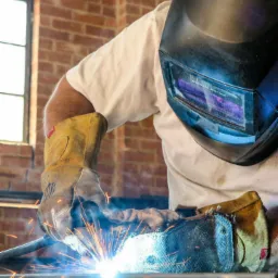 Osoba w masce spawalniczej i rękawicach ochronnych podczas spawania metalu, widoczne iskry i płomienie, tło z ceglaną ścianą i oknem.