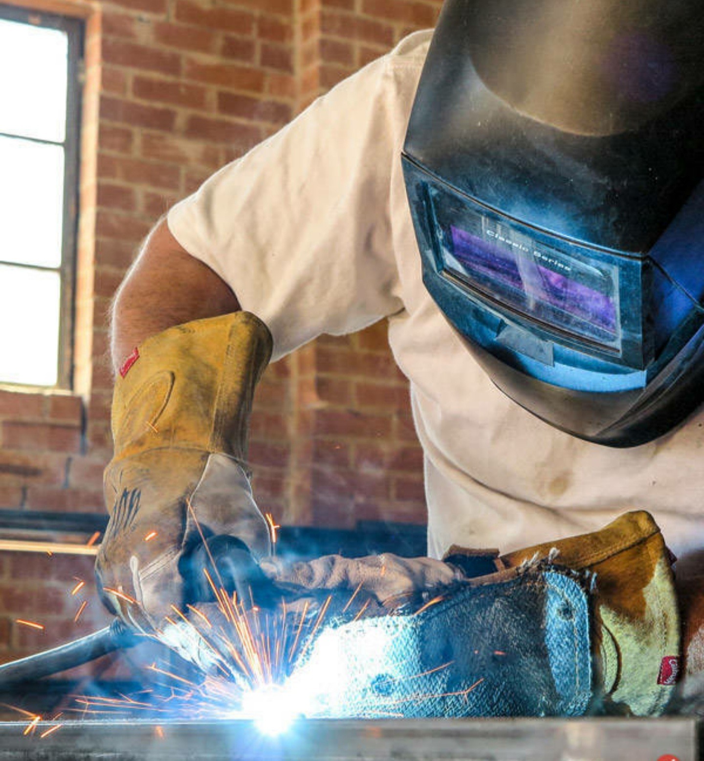 Osoba w masce spawalniczej i rękawicach ochronnych podczas spawania metalu, widoczne iskry i płomienie, tło z ceglaną ścianą i oknem.