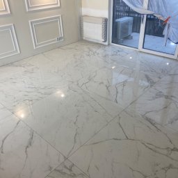 DexoBau Karwowski Damian - Błyszcząca podłoga z płytek imitujących biały marmur z szarymi żyłami, świeżo położona w pomieszczeniu z dekoracyjną sztukaterią na ścianach i oknem balkonowym zabezpieczonym folią.