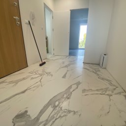 DexoBau Karwowski Damian - Nowe, białe płytki podłogowe imitujące marmur z wyraźnym szarym wzorem w przestronnym, jasnym przedpokoju z widokiem na balkon i stojącą miotłą.