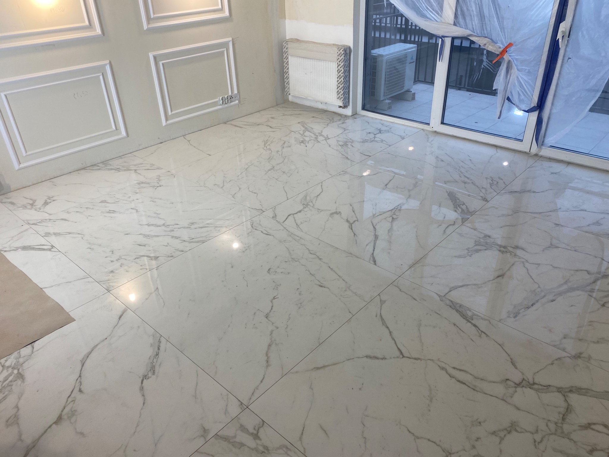Błyszcząca podłoga z płytek imitujących biały marmur z szarymi żyłami, świeżo położona w pomieszczeniu z dekoracyjną sztukaterią na ścianach i oknem balkonowym zabezpieczonym folią.