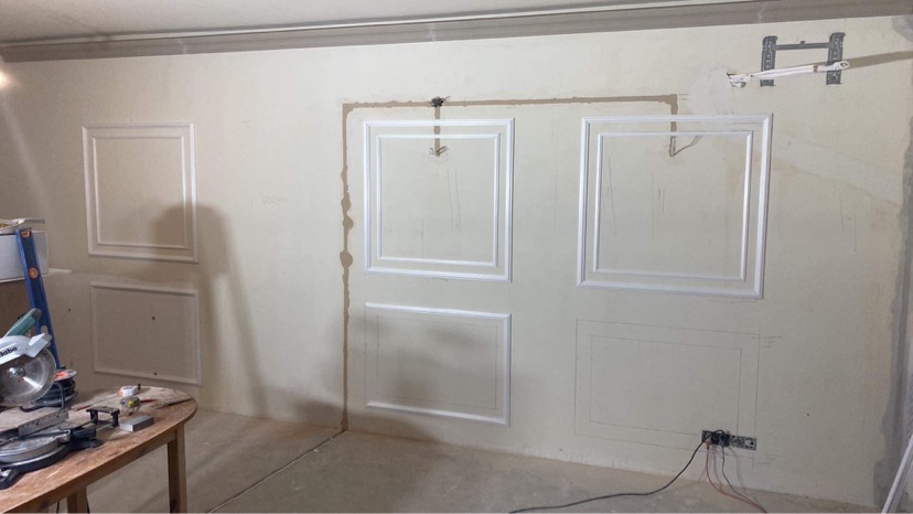 Ściana w trakcie dekorowania sztukaterią w formie kwadratowych ramek, widoczne ślady po ołówku i niedoskonałości na ścianie, na pierwszym planie stół z piłą ukośnicą.