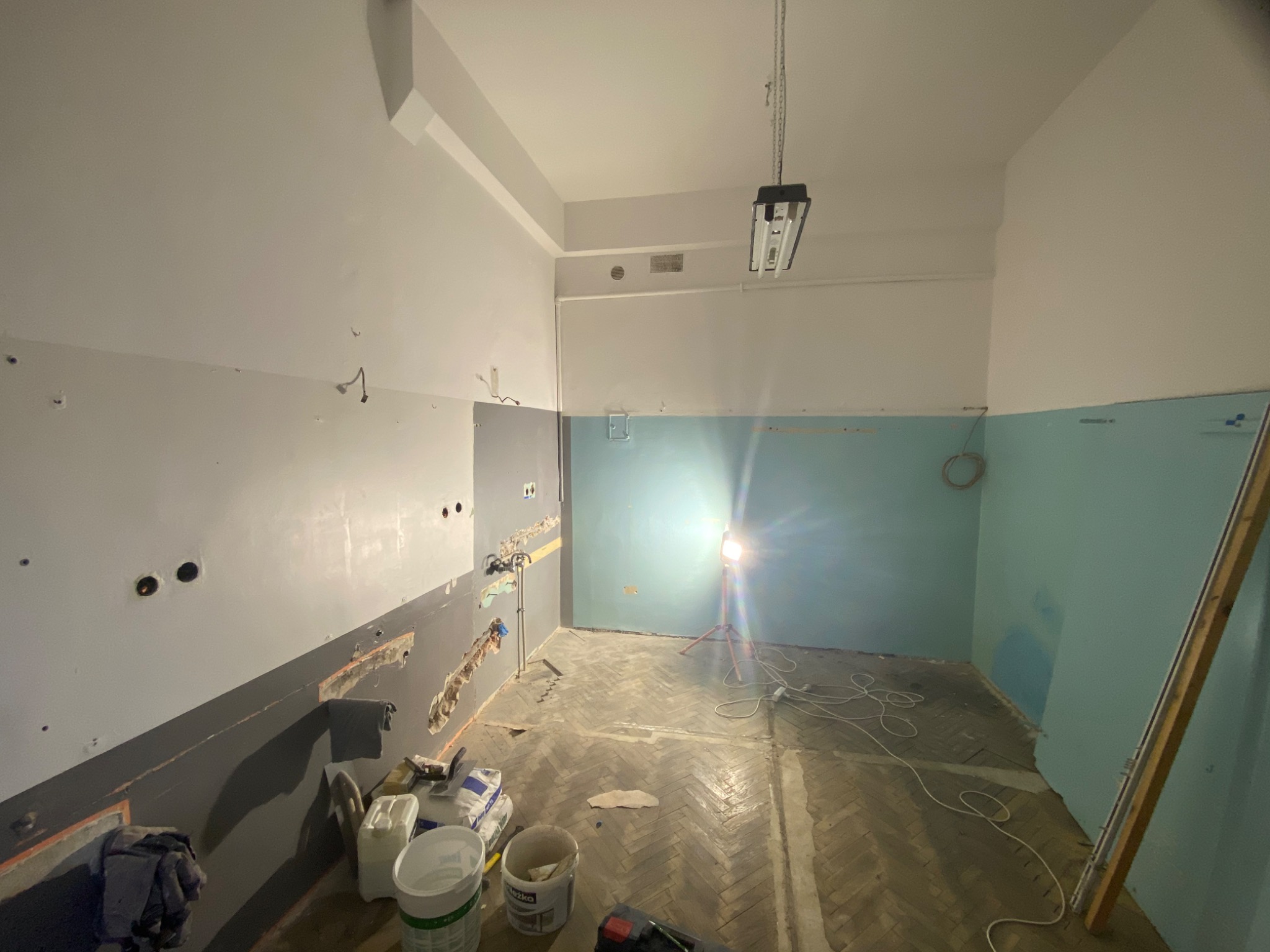 Pokój w trakcie remontu: ściany częściowo pokryte szpachlą, widoczne puszki elektryczne, podłoga parkietowa, narzędzia i materiały budowlane w dolnej części kadru, lampa budowlana na trójnogu...