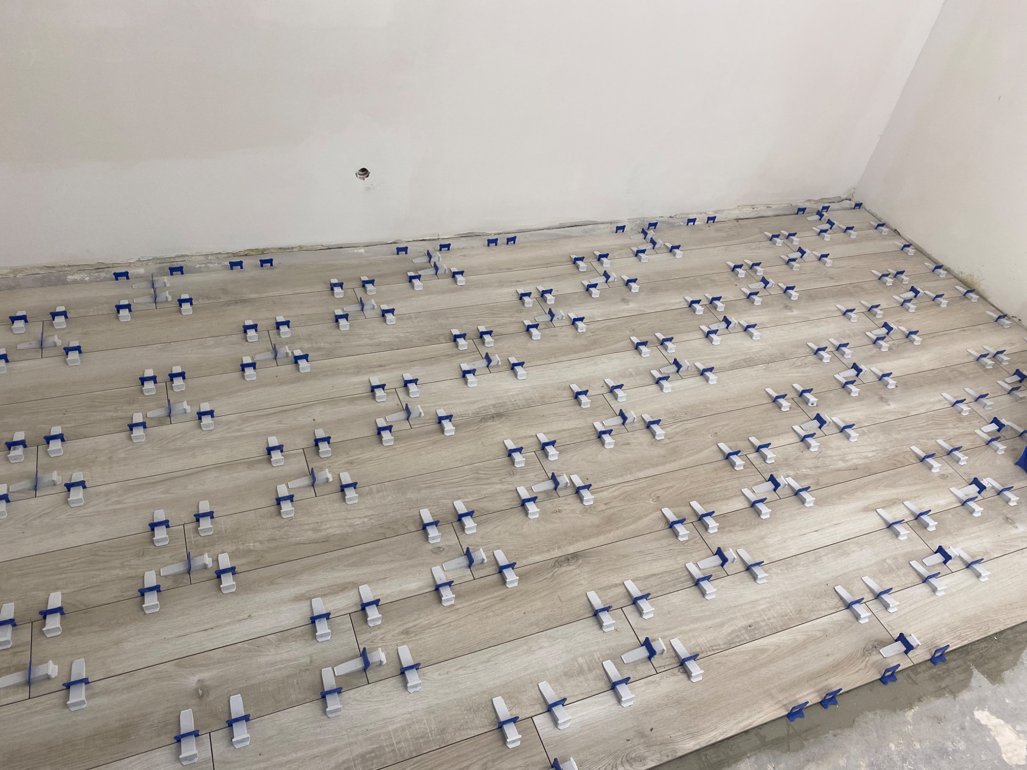 Podłoga w trakcie układania płytek imitujących drewno, z widocznymi niebiesko-białymi klipsami poziomującymi, ściany przygotowane do dalszych prac.