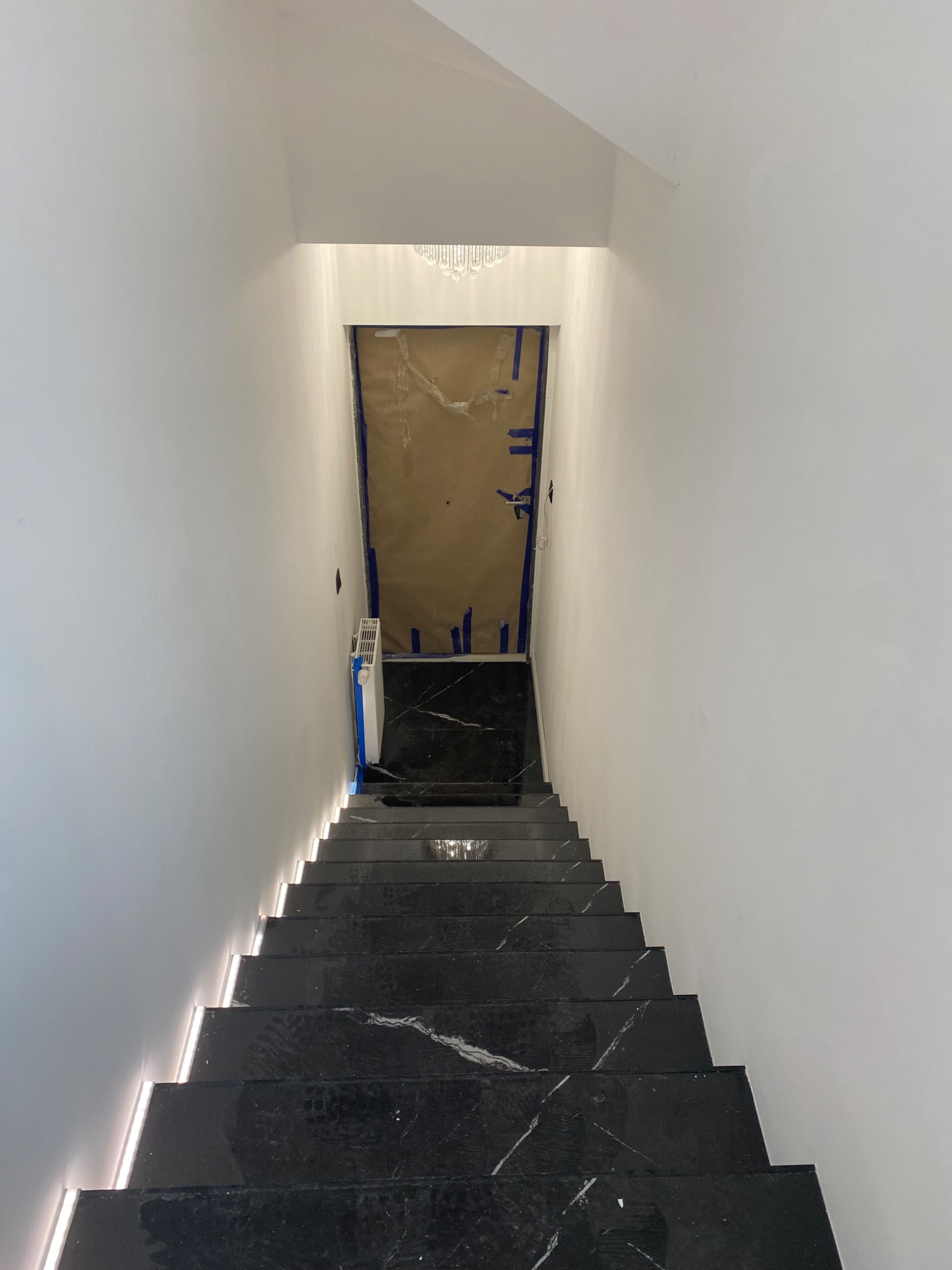 Widok z góry na czarne marmurowe schody z podświetleniem LED, prowadzące w dół do pomieszczenia z zabezpieczonymi drzwiami i widocznym fragmentem żyrandola.
