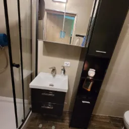 Nowoczesna łazienka z kabiną prysznicową z czarnymi ramami, umywalką z szafką w kolorze czarnym, wysoką szafką z półkami na dekoracje i płytkami imitującymi marmur na podłodze.