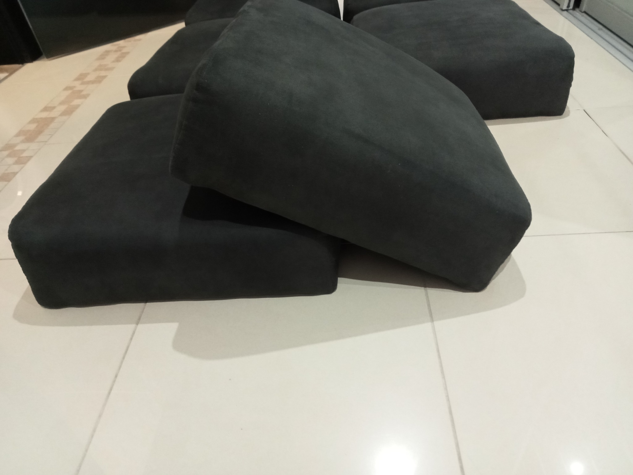 Ciemnoszare, tapicerowane poduszki siedziskowe na jasnej, wzorzystej podłodze. Widoczne przeszycia i zaokrąglone krawędzie. Ujęcie z góry, lekko z ukosa.