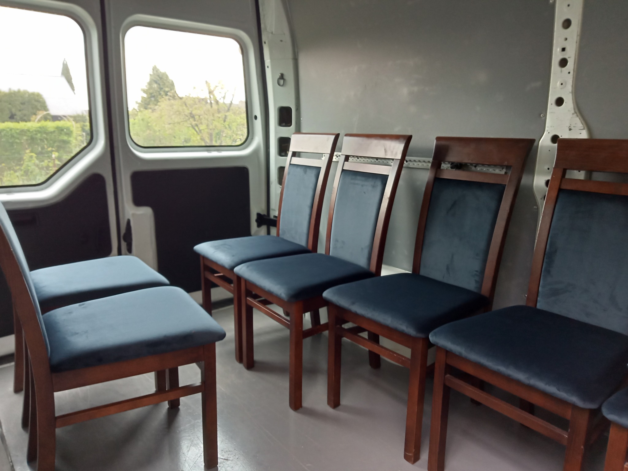 Krzesła z ciemnoniebieską tapicerką w przestrzeni ładunkowej dostawczaka. Widok z boku, oświetlenie dzienne, widoczne elementy konstrukcyjne pojazdu.