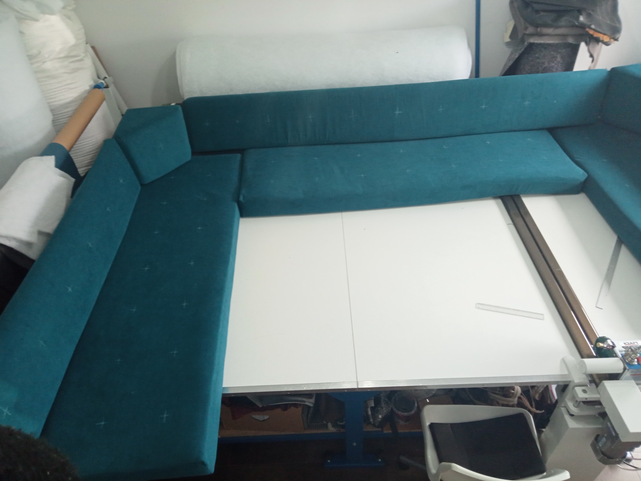 Turkusowy narożnik w trakcie tworzenia, widoczne szpilki na materiale. Narożnik częściowo ułożony na białym stole, w tle materiały i narzędzia tapicerskie.