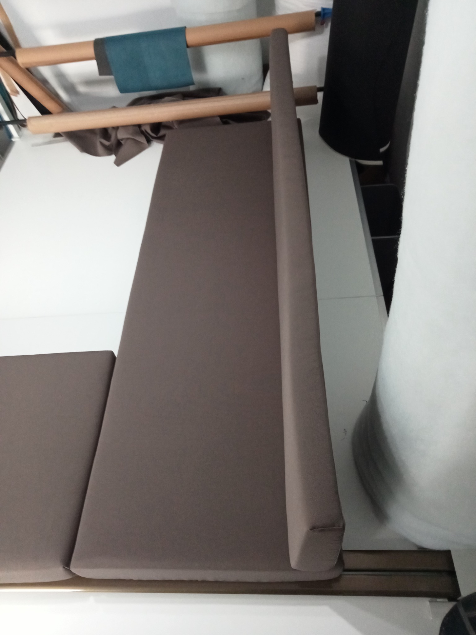 Tapicerowany, brązowy zagłówek i siedzisko sofy w warsztacie. Widoczne rolki materiałów i narzędzia. Ukończony element mebla przed montażem.