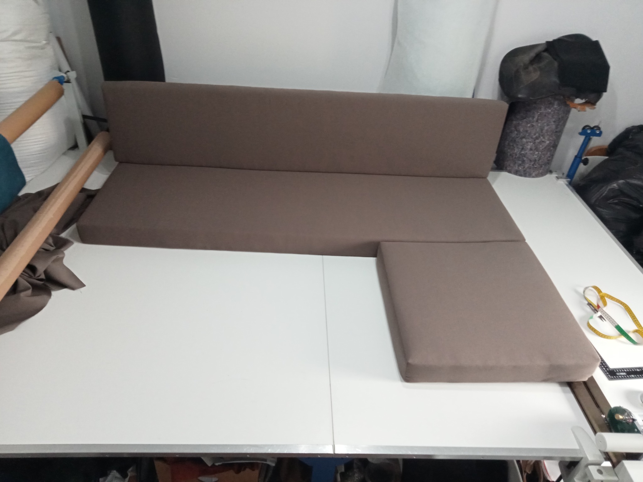 Brązowe, tapicerowane elementy sofy narożnej, w trakcie produkcji, leżą na białym stole. Widoczne materiały i miarka krawiecka. Ujęcie z góry, w warsztacie.