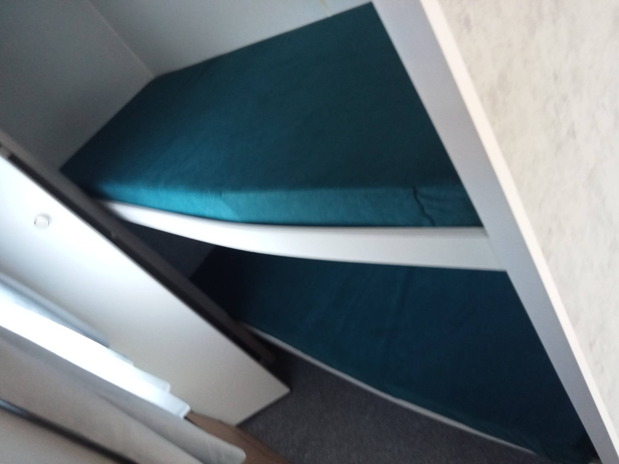 Turkusowy materac na białym, drewnianym stelażu łóżka piętrowego, widziany z dołu pod kątem. Szara wykładzina dywanowa i biała ściana w tle.