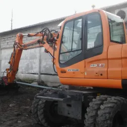Pomarańczowa koparka kołowa New Holland MH3.6 na placu budowy, z widocznym numerem telefonu 503 775 191, pracująca przy wykopie w pobliżu betonowego budynku z klimatyzatorami.