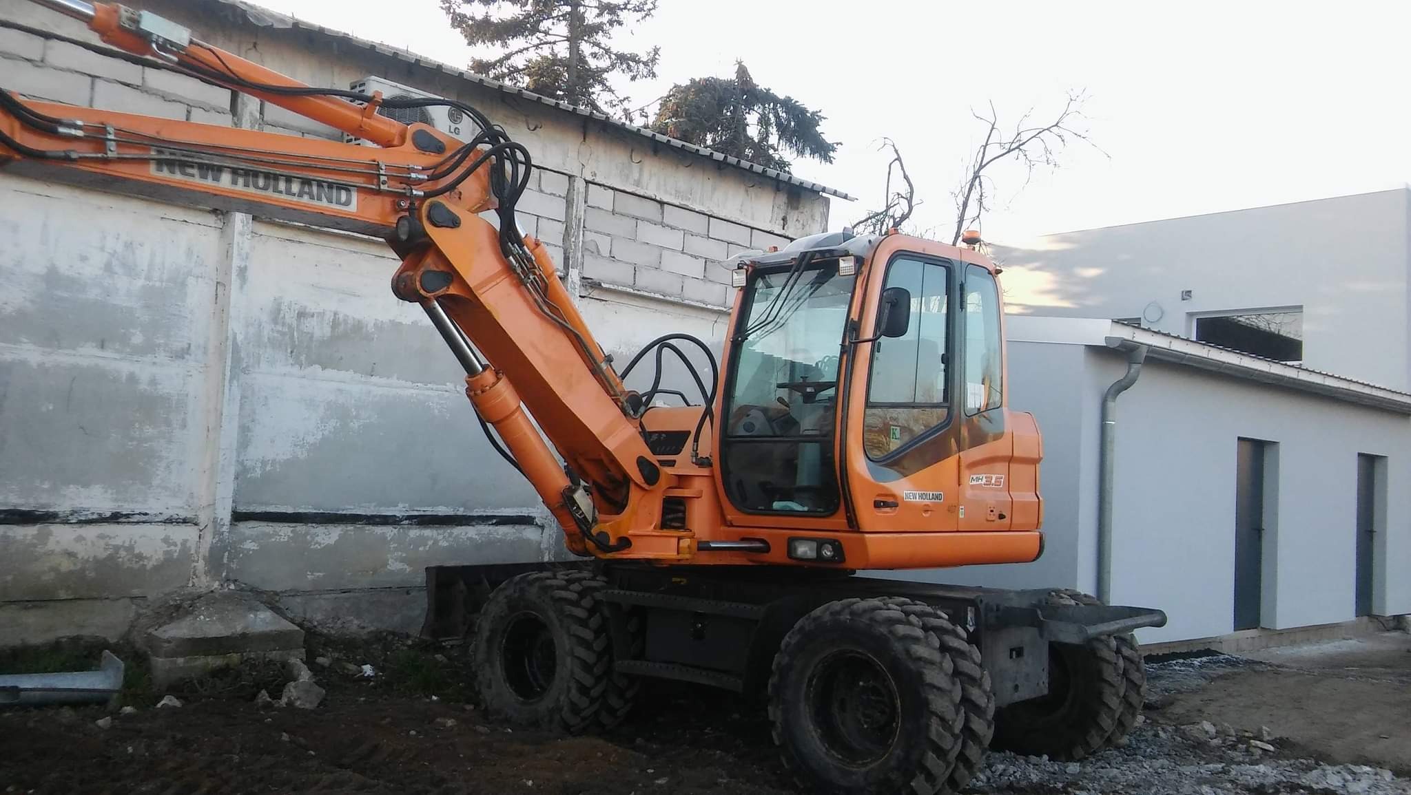 Pomarańczowa koparka kołowa New Holland MH3.6 zaparkowana przy betonowym murze i nowo wybudowanym budynku z szarą elewacją.