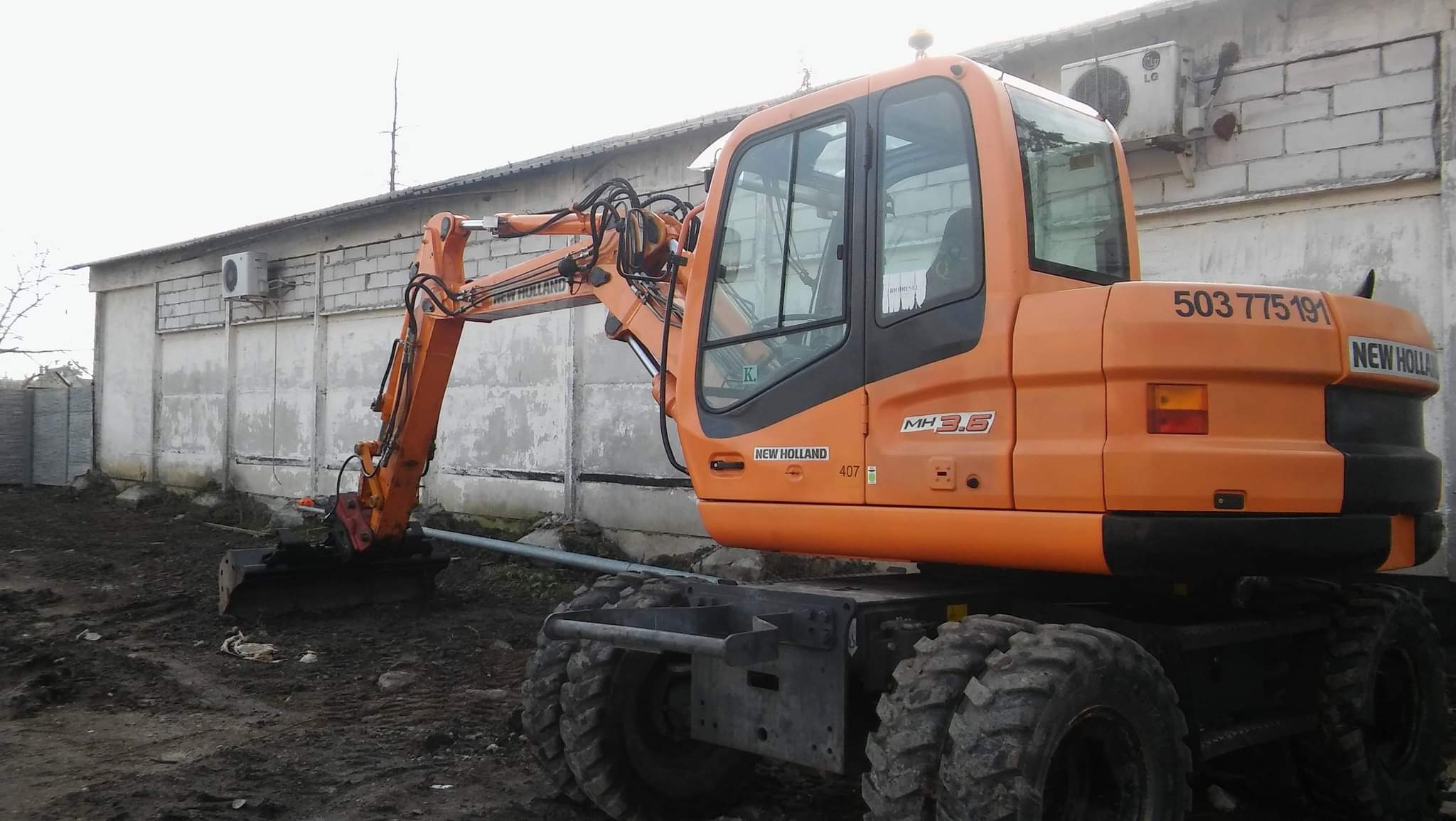 Pomarańczowa koparka kołowa New Holland MH3.6 na placu budowy, z widocznym numerem telefonu 503 775 191, pracująca przy wykopie w pobliżu betonowego budynku z klimatyzatorami.