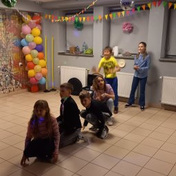 MK'smile Krzysztof Sobik - Dzieci bawią się w rzędzie w udekorowanym pomieszczeniu z balonami, girlandami i dyskotekową kulą. Jedna dziewczynka stoi, reszta w pozycji kucznej lub w ruchu.