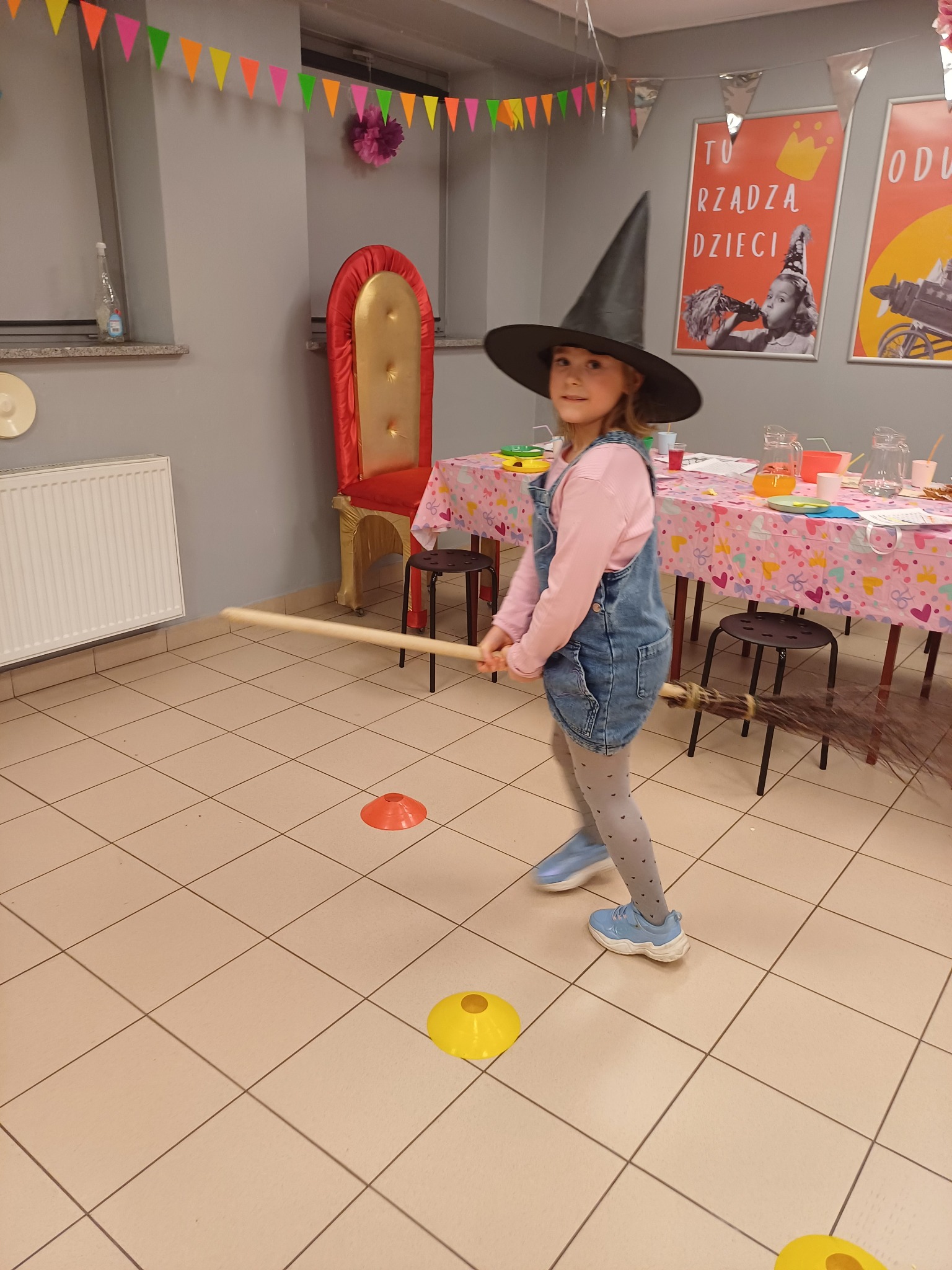 Dziewczynka w stroju czarownicy z miotłą, na tle sali z dekoracjami urodzinowymi i stołem z napojami. Konkurs zręcznościowy na imprezie dla dzieci.