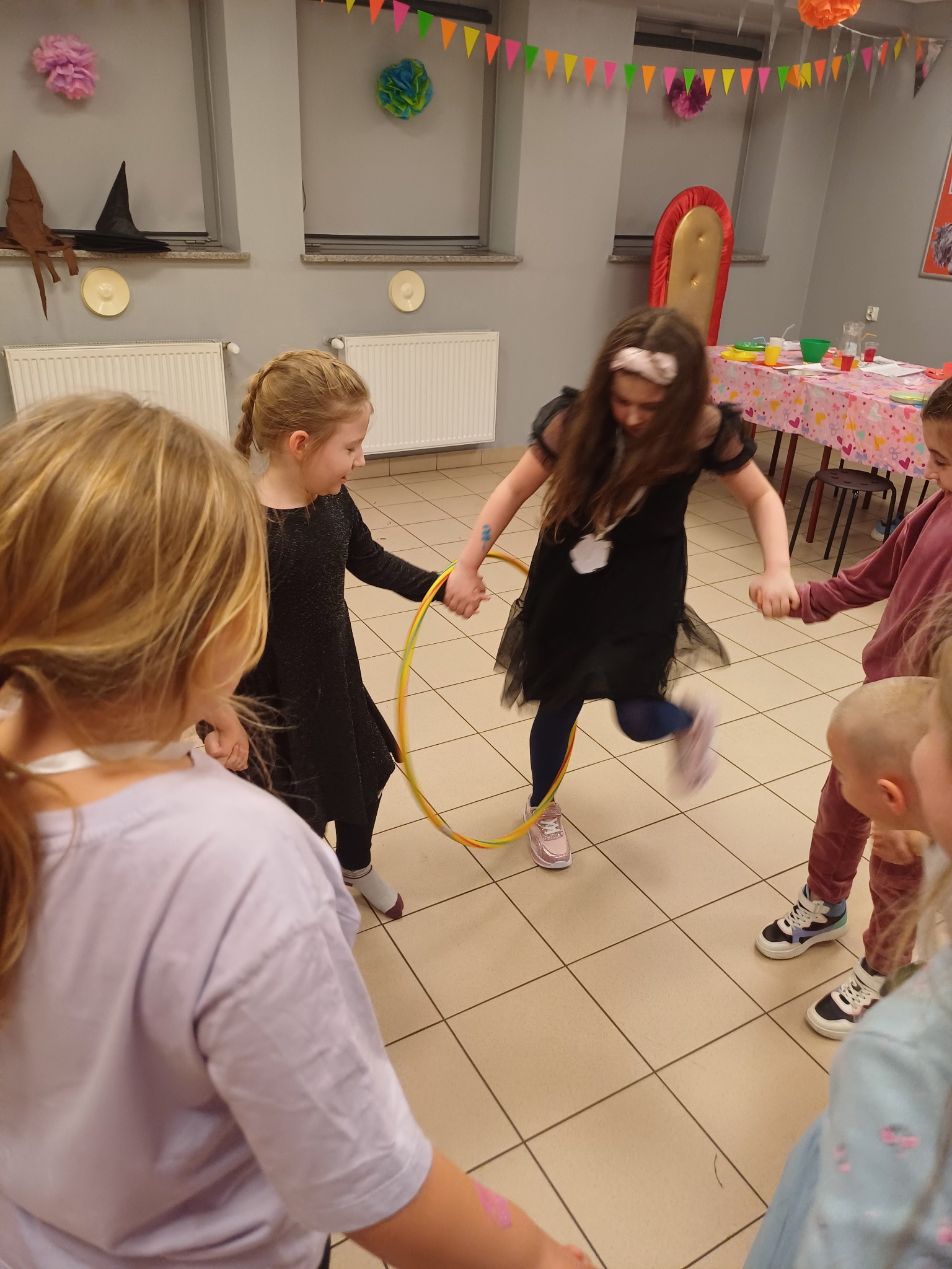 Dzieci bawią się hula-hoop podczas imprezy urodzinowej. Dekoracje wiszą na ścianie, a stół z przekąskami stoi w tle. Dziewczynka w czarnej sukience skacze przez obręcz.