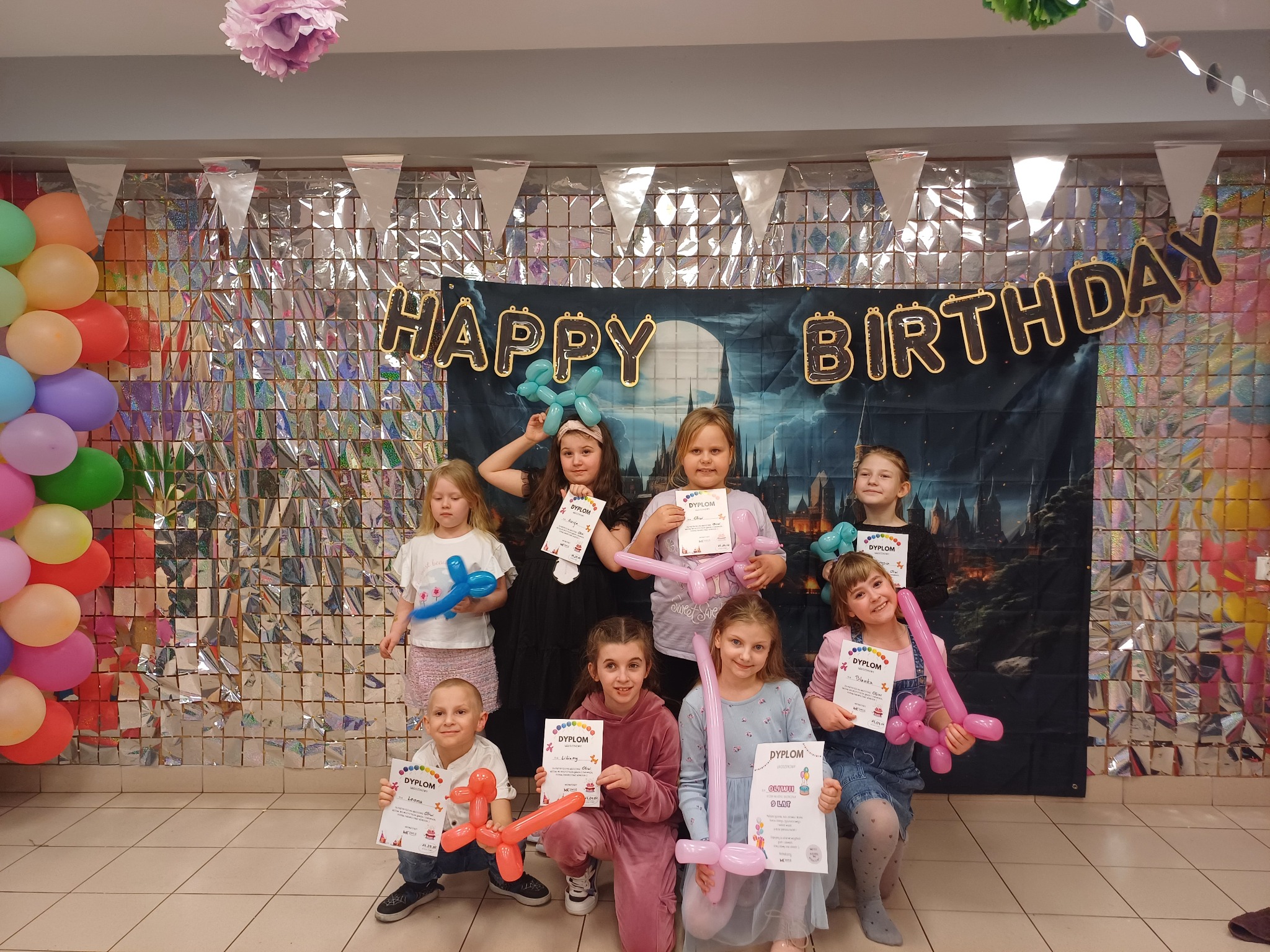 Uśmiechnięte dzieci z balonami i dyplomami na tle dekoracji urodzinowych. Balony w kształcie zwierząt i napis 'Happy Birthday'.