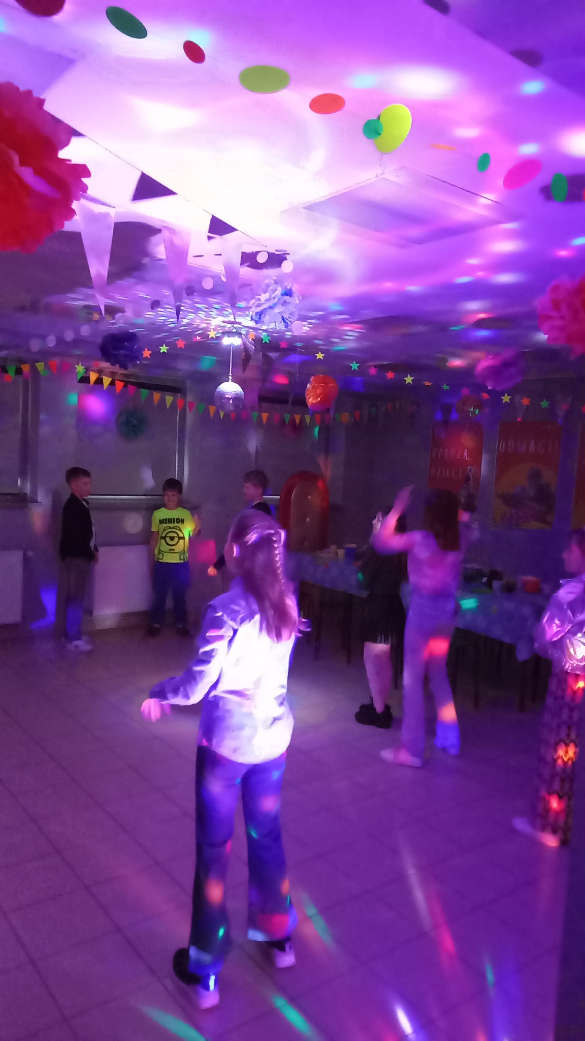 Urodziny dziecka w sali z dekoracjami: kolorowe girlandy, balony, światła dyskotekowe. Dzieci bawią się w rytm muzyki. Na ścianach plakaty. Atmosfera radosna i pełna energii.