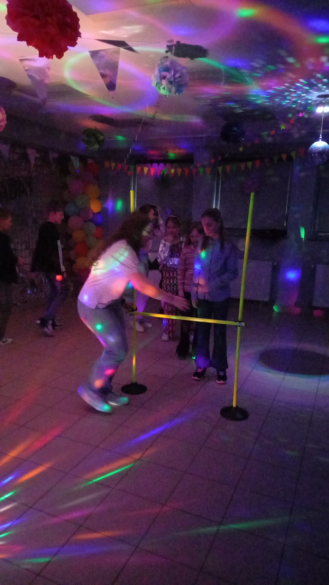 Dziewczyna w limbo pod kolorowymi światłami na imprezie dla dzieci. W tle grupa dzieci i dekoracje, tworzące radosną atmosferę zabawy.