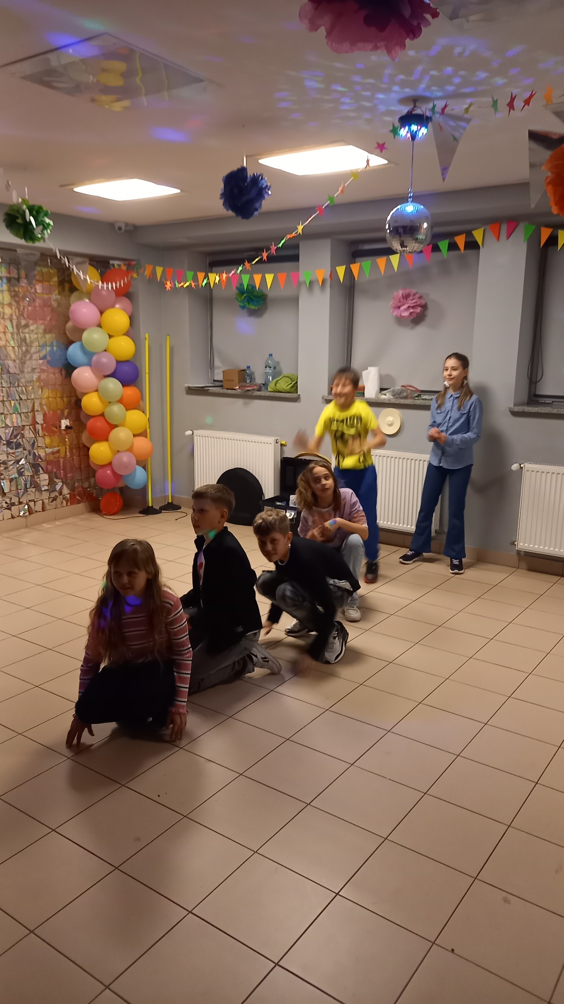 Dzieci bawią się w rzędzie w udekorowanym pomieszczeniu z balonami, girlandami i dyskotekową kulą. Jedna dziewczynka stoi, reszta w pozycji kucznej lub w ruchu.