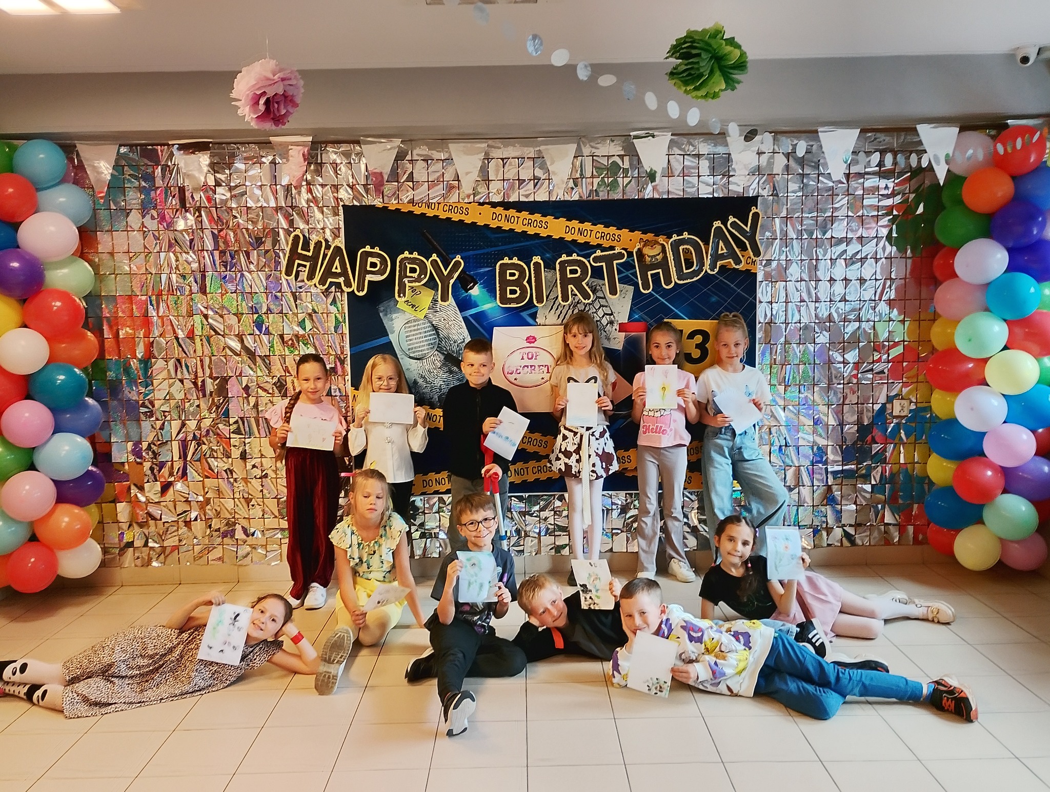 Grupa dzieci na tle dekoracji urodzinowych z motywem detektywistycznym, trzymających rysunki. Balony po bokach, błyszcząca ściana w tle. Uśmiechnięte twarze.