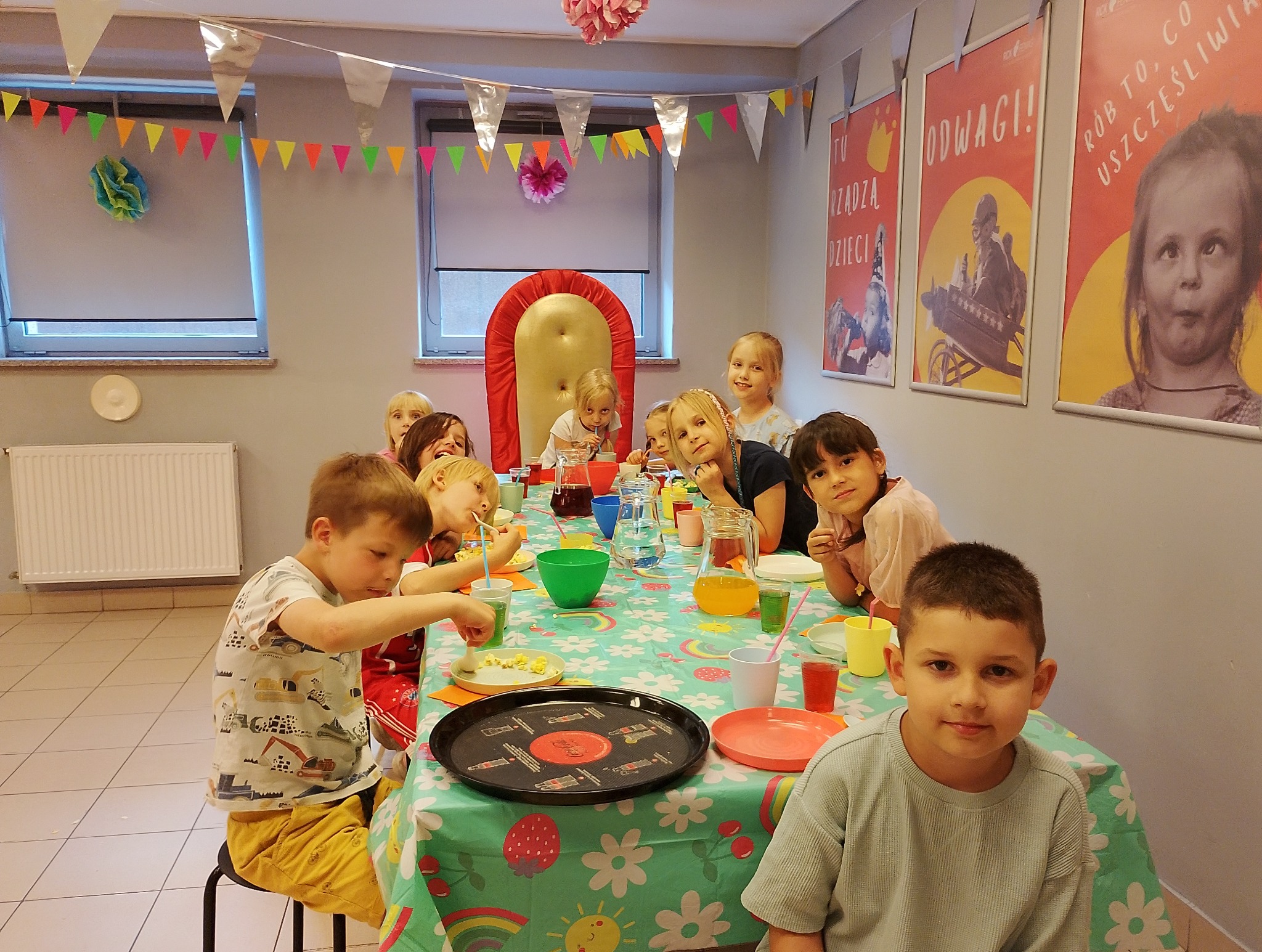 Urodziny dziecka: grupa dzieci siedzi przy stole z kolorowym obrusem, jedzą i piją. W tle dekoracje urodzinowe i plakaty.