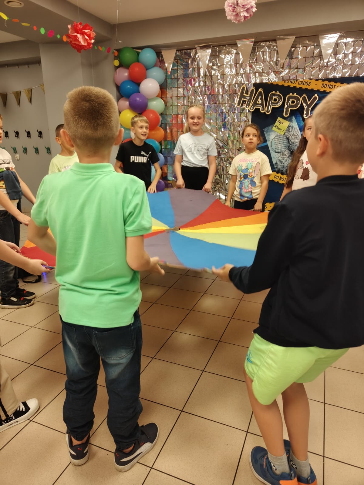 Grupa dzieci bawi się kolorową chustą animacyjną w sali udekorowanej balonami i napisem 'Happy'. Widoczne elementy dekoracyjne i radosne miny uczestników zabawy.