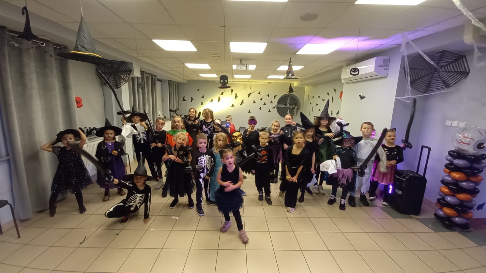 Grupa dzieci w kostiumach Halloween, w tym czarownice i szkielety, pozuje w udekorowanym pokoju z balonami i dekoracjami tematycznymi. Widoczne rekwizyty i kostiumy.