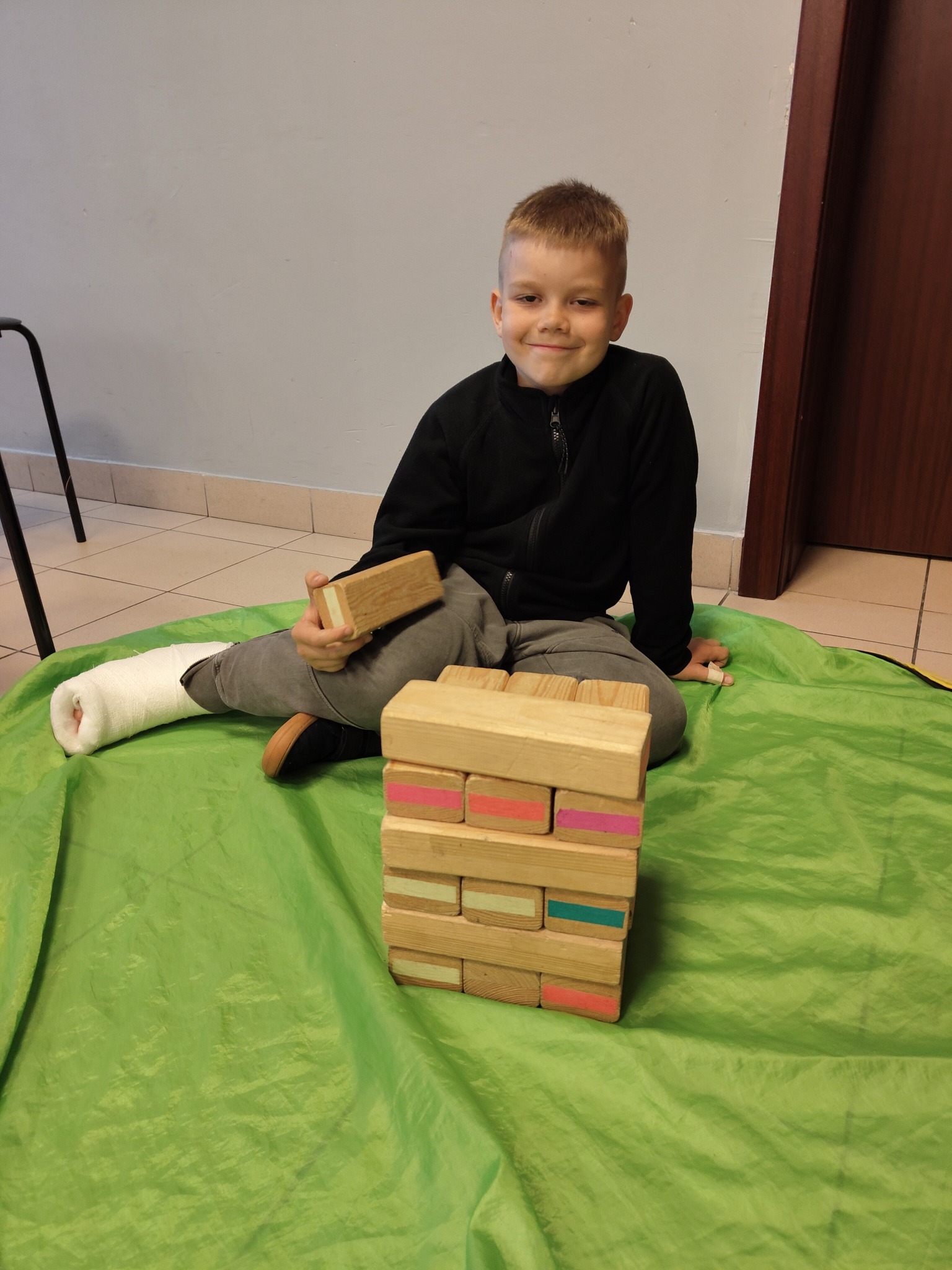 Chłopiec z nogą w gipsie siedzi na zielonej macie i gra w drewnianą wieżę Jenga z kolorowymi elementami. Uśmiechnięty, w czarnej bluzie.