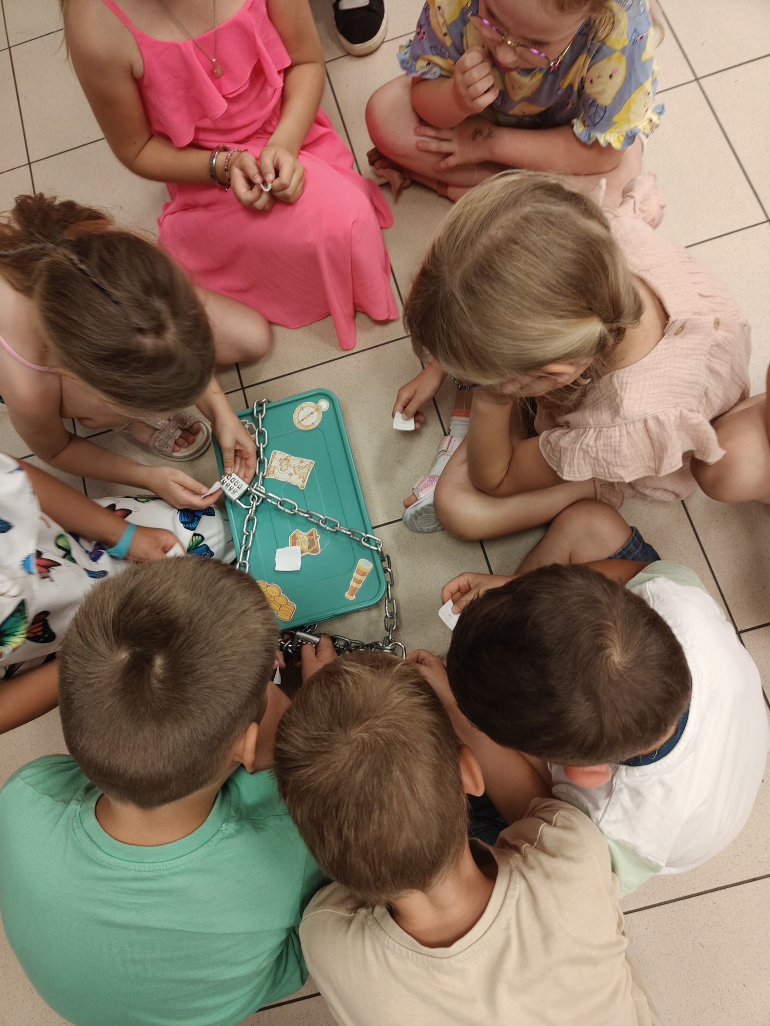 Dzieci siedzą w kręgu na podłodze, rozwiązując zagadkę z zamkniętą na łańcuch skrzynką z naklejkami map i skarbów, trzymając w rękach kartki z kodami.