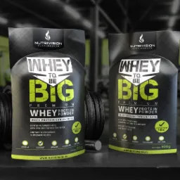 Dwa opakowania czarnego proszku proteinowego 'Whey to be Big' z logiem Nutrivision Supplements, obok hantli na tle siłowni.