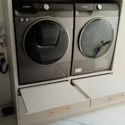 Szaro-beżowa zabudowa z pralką i suszarką Samsung, szuflady z poziomymi, złotymi uchwytami, marmurowa podłoga.