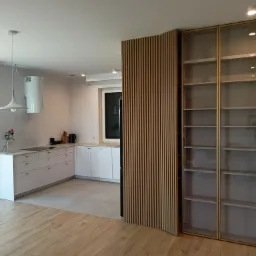 Nowoczesna kuchnia z białymi szafkami, drewnianą ścianką działową z pionowych listew i przeszkloną szafą z półkami, widok na jasne drewniane podłogi.