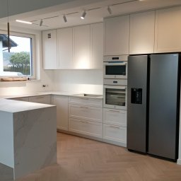 Stablo Meble - Jasna kuchnia z wyspą i marmurowym blatem. Białe szafki z złotymi uchwytami, sprzęt AGD w zabudowie. Parkiet ułożony w jodełkę. Nowoczesne oświetlenie.