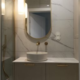 Stablo Meble - Elegancka łazienka z owalnym lustrem w złotej ramie, umywalką nablatową i szafką pod umywalką z subtelnym oświetleniem LED. Marmurowe płytki na ścianach i podłodze.