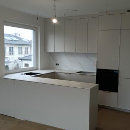 Stablo Meble - Nowoczesna, jasna kuchnia z wyspą i marmurowym blatem. Minimalistyczne szafki bez uchwytów, okno z widokiem na budynek. Oświetlenie punktowe podkreśla detale.